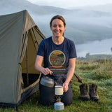 Woman in the Adventure Before Dementia Camping T-Shirt in Navy pouring a brew from a camping stove outside her tent at dawn
