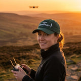 Woman wearing the Task "Drone Pilot" cap in Spruce, flying a drone over moorland at sunset