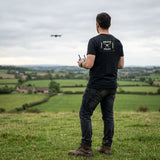 Man flying a drone on a hilltop in the English countryside, wearing the Vintage Black Drone Pilot T-shirt with the large stencil patch print on the back