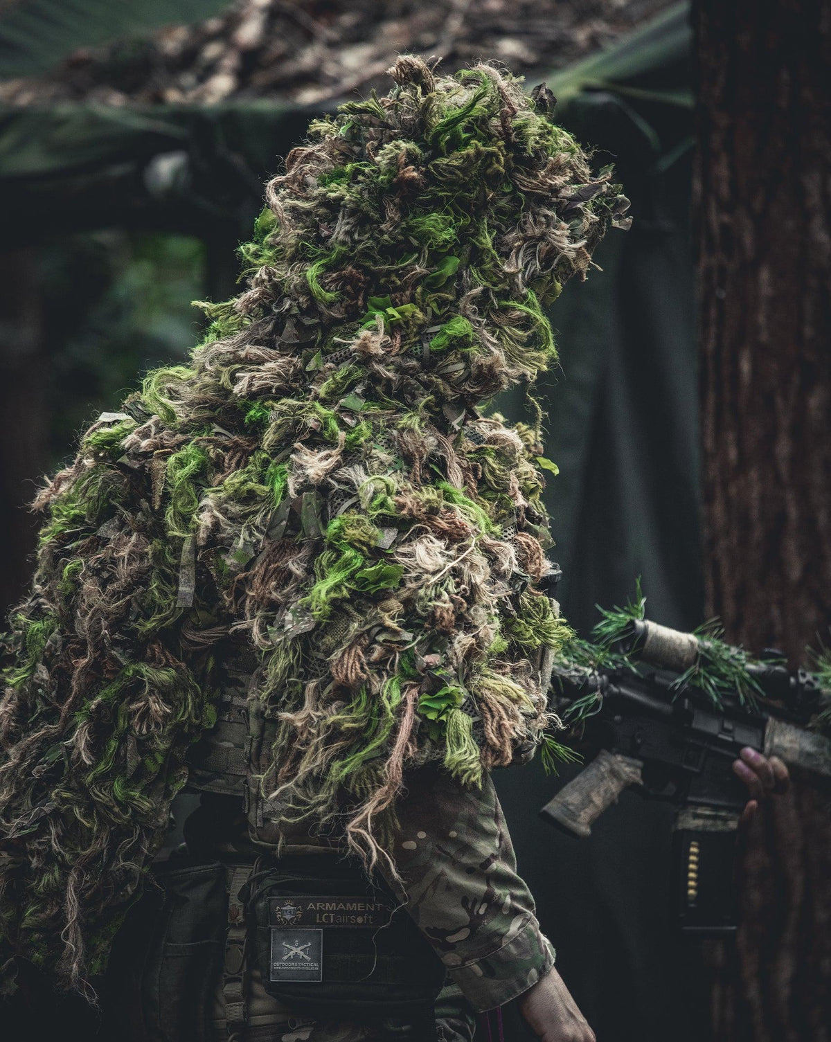 Zero Alpha Designs mid cape ghillie suit in the bright green Summer scheme, worn over a plate carrier with a camouflaged rifle in dark woodland