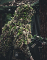 Zero Alpha Designs mid cape ghillie suit in the bright green Summer scheme, worn over a plate carrier with a camouflaged rifle in dark woodland