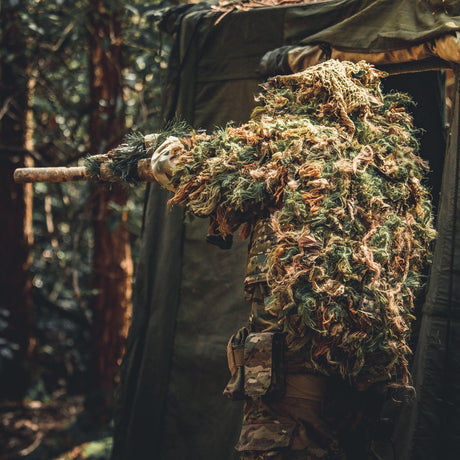 Shooter in a Zero Alpha Designs full cape ghillie suit shouldering a wrapped rifle from the mouth of a woodland hide