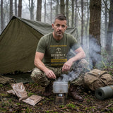 Man in Military Green "In My Defence, I Was Left Unsupervised" T-shirt crouched over a smoking mess tin at a woodland basha