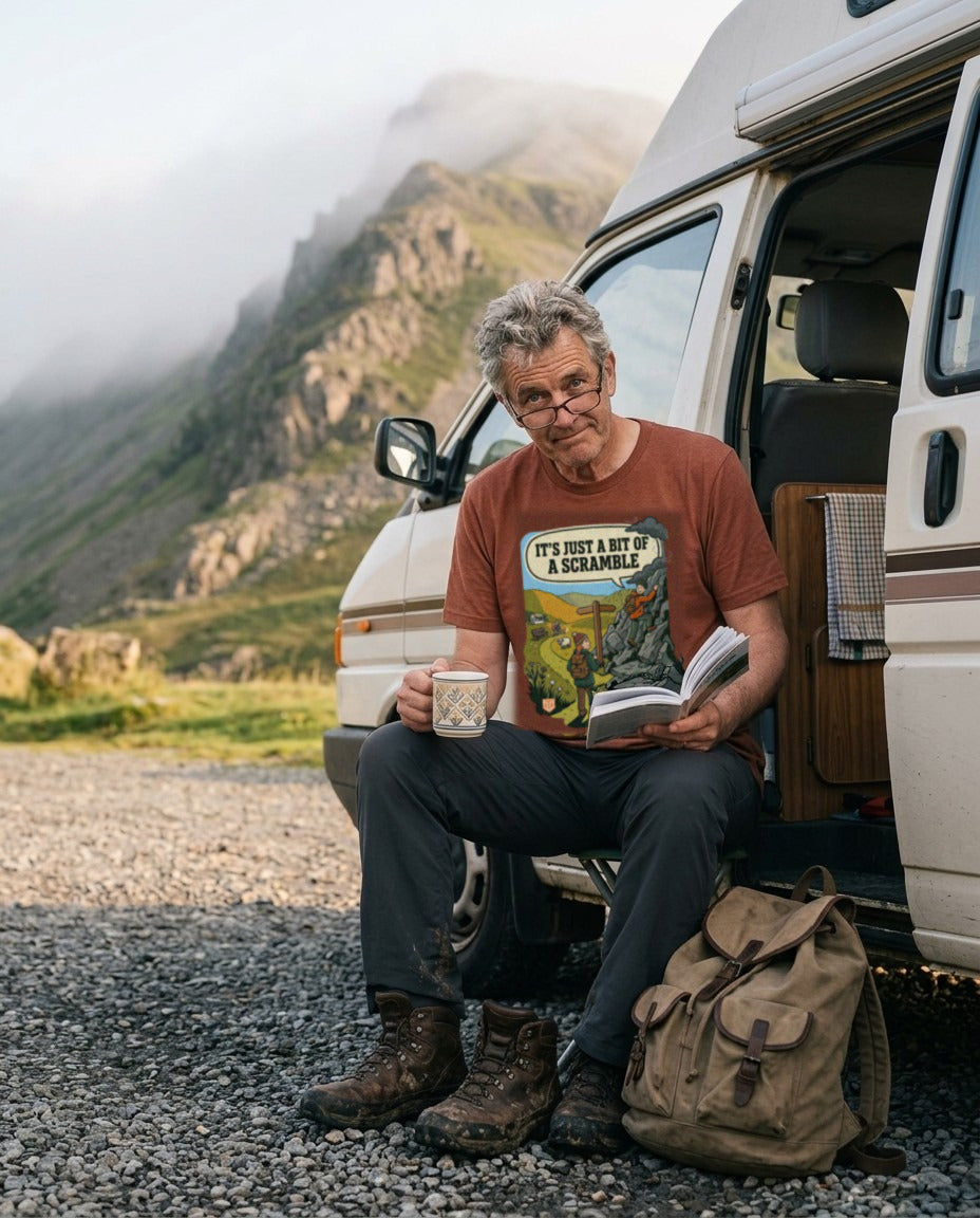 Man in the It's Just a Bit of a Scramble T-Shirt in Heather Clay with a mug of tea and a guidebook in a campervan doorway below a misty ridge