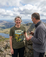 Woman in the It's Just a Bit of a Scramble T-Shirt in Military Green laughing at a summit cairn while her friend holds a flask cup