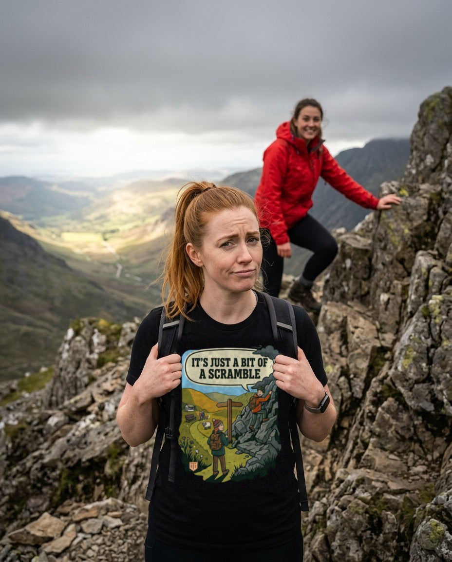 Walker in the It's Just a Bit of a Scramble T-Shirt in Vintage Black looking doubtful at the foot of a rocky ridge while her friend grins from the rocks above