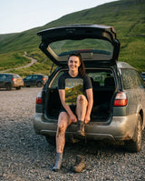 Woman in the It's Just Over the Brow T-Shirt in Dark Grey Heather sitting in the open boot of a muddy car after a hill walk