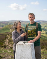 Couple at a trig point on a hill summit, the man wearing the It's Just Over the Brow T-Shirt in Forest