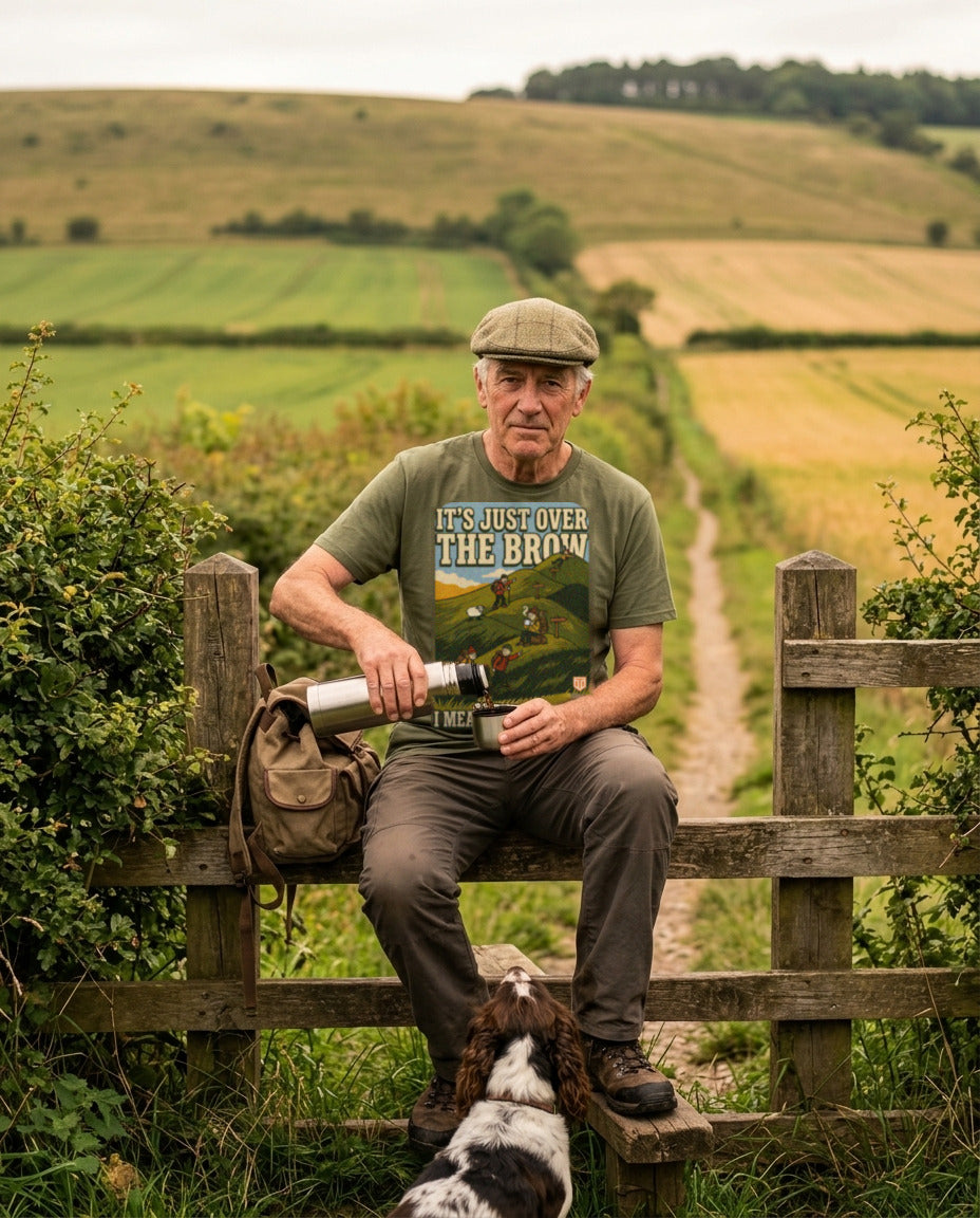 Walker in a flat cap on a stile pouring tea from a flask, wearing the It's Just Over the Brow T-Shirt in Military Green, with a spaniel at his feet