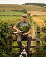 Walker in a flat cap on a stile pouring tea from a flask, wearing the It's Just Over the Brow T-Shirt in Military Green, with a spaniel at his feet