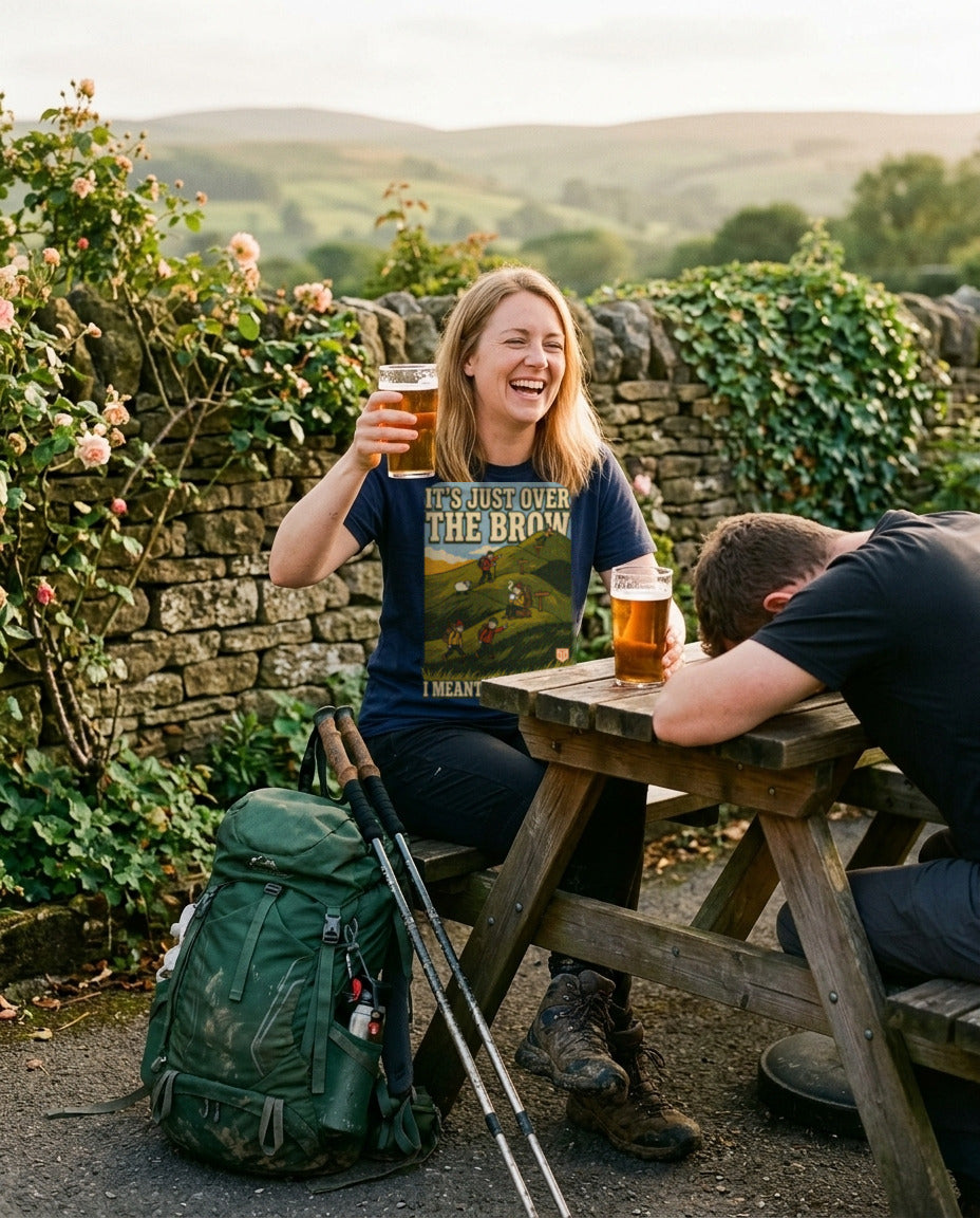 Woman in the It's Just Over the Brow T-Shirt in Navy raising a pint in a pub garden after a walk while her friend slumps on the table