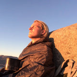 Camper wrapped in the Olive Green Snugpak Jungle Blanket WGTE resting against a rock with a camp mug at sunset