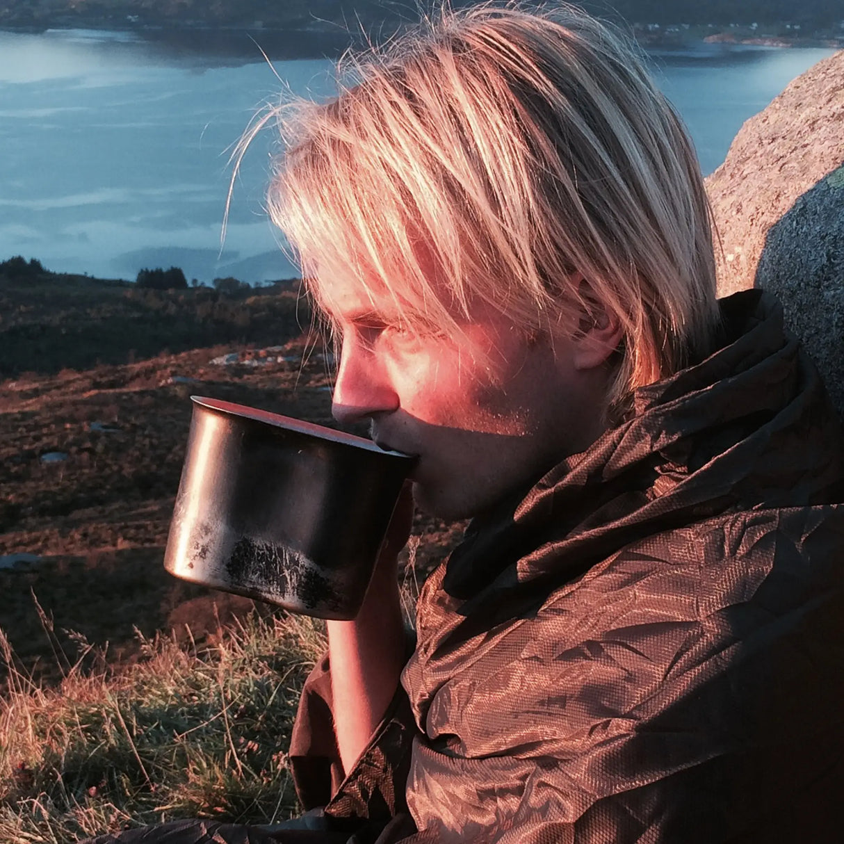 Camper wrapped in the Olive Green Snugpak Jungle Blanket WGTE drinking from a metal camp mug at a mountain viewpoint