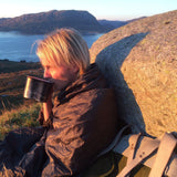 Camper wrapped in the Snugpak Jungle Blanket WGTE in Olive Green leaning on a rock with a camp mug above a fjord