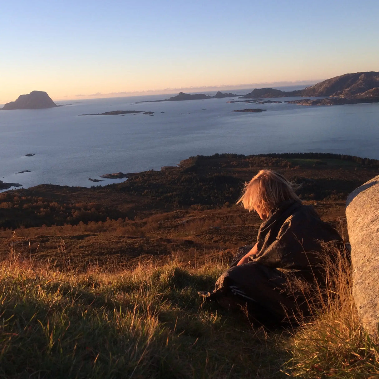 Camper wrapped in the Olive Green Snugpak Jungle Blanket WGTE sitting on a grassy clifftop overlooking a fjord at sunset