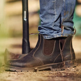 Close-up of brown Mongrel K9 dealer boots worn with jeans on a dusty farmyard track