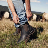 Man in a grassy field tucking his jeans into brown Mongrel K9 dealer boots, sheep and hay bales behind