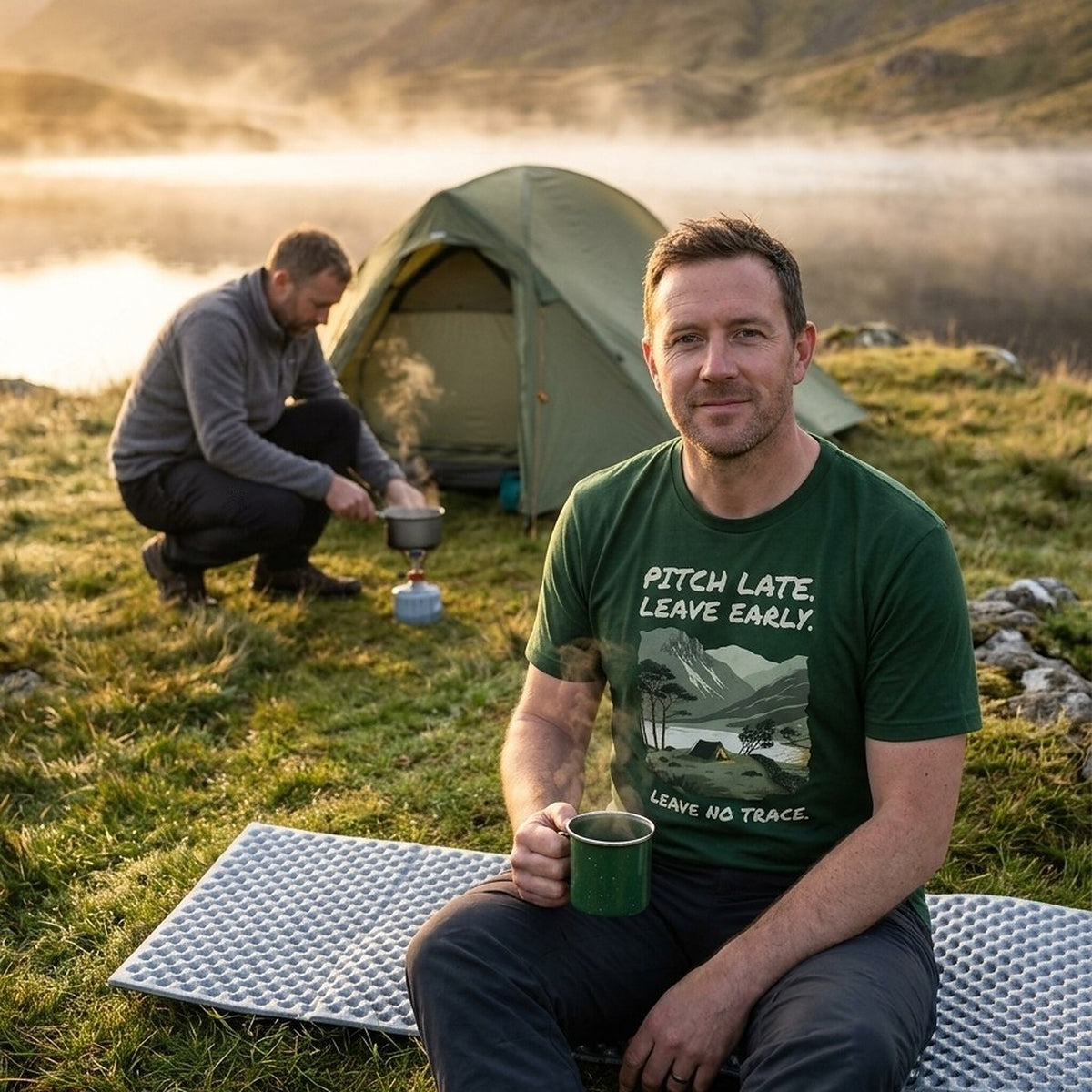 Man with an enamel mug of tea by a misty tarn at sunrise, wearing the Pitch Late Leave Early T-Shirt in Forest, while his friend boils water on a gas stove