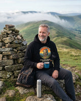 Walker with a mug of tea beside a summit cairn wearing the Sloth Hiking Club: Hilltop Hoodie in Black