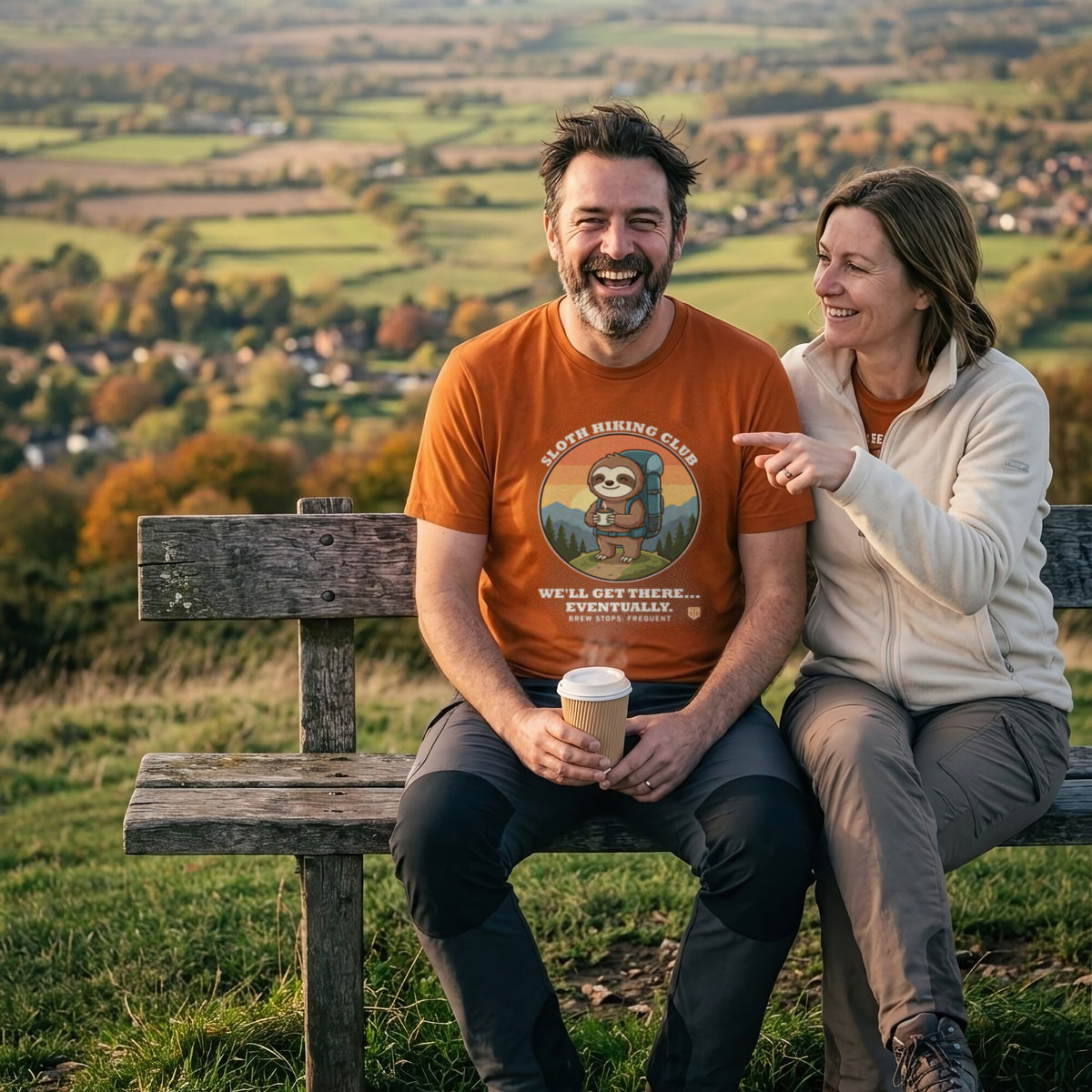 Couple laughing on a hilltop bench above autumn fields, the man wearing the Sloth Hiking Club: Hilltop T-Shirt in Autumn