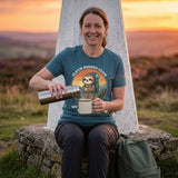 Woman pouring tea from a flask at a trig point at sunset, wearing the Sloth Hiking Club: Hilltop T-Shirt in Heather Deep Teal