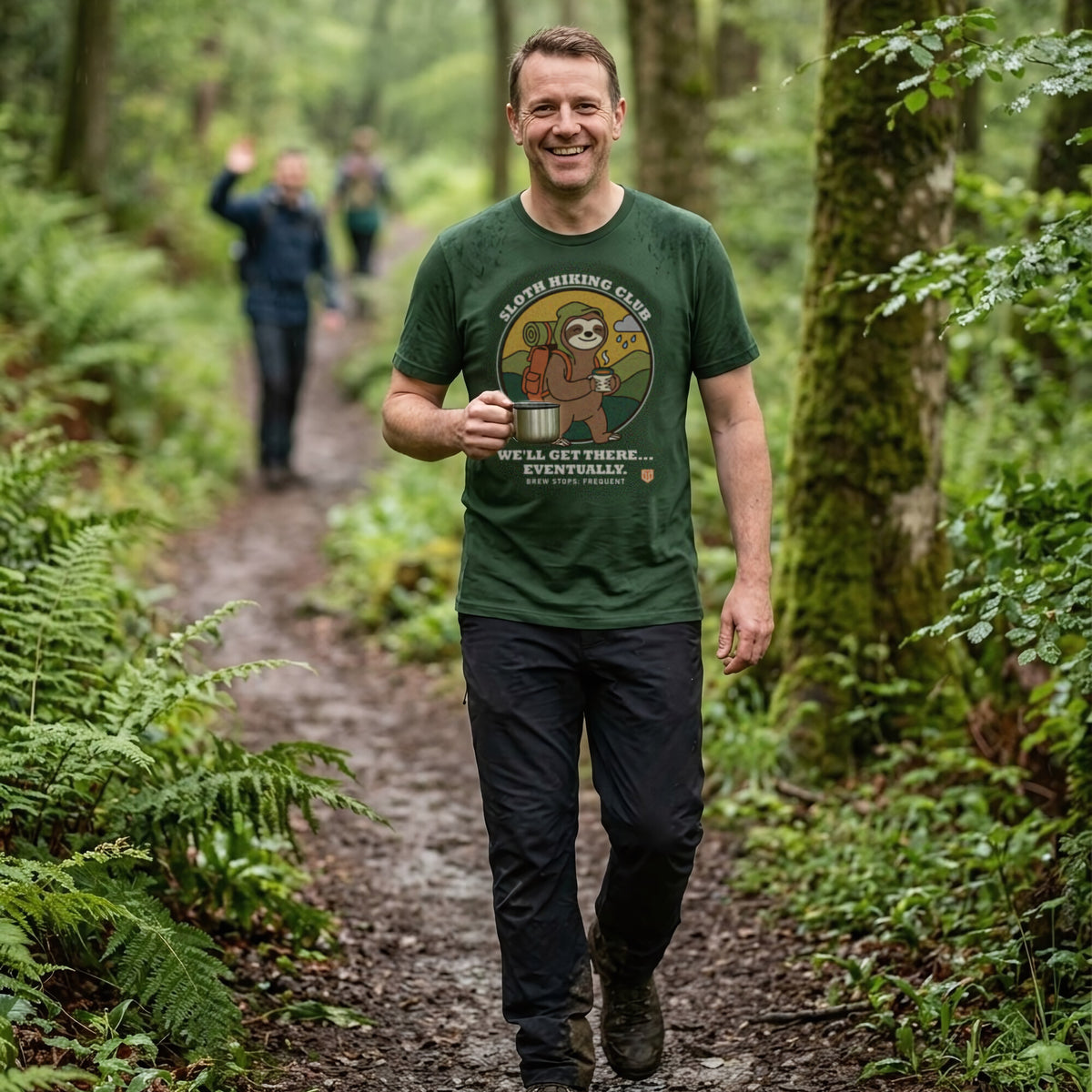 Man with a steaming mug walking a muddy woodland path in light rain, friends waving ahead, wearing the Sloth Hiking Club: Walking T-Shirt in Forest