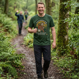Man with a steaming mug walking a muddy woodland path in light rain, friends waving ahead, wearing the Sloth Hiking Club: Walking T-Shirt in Forest
