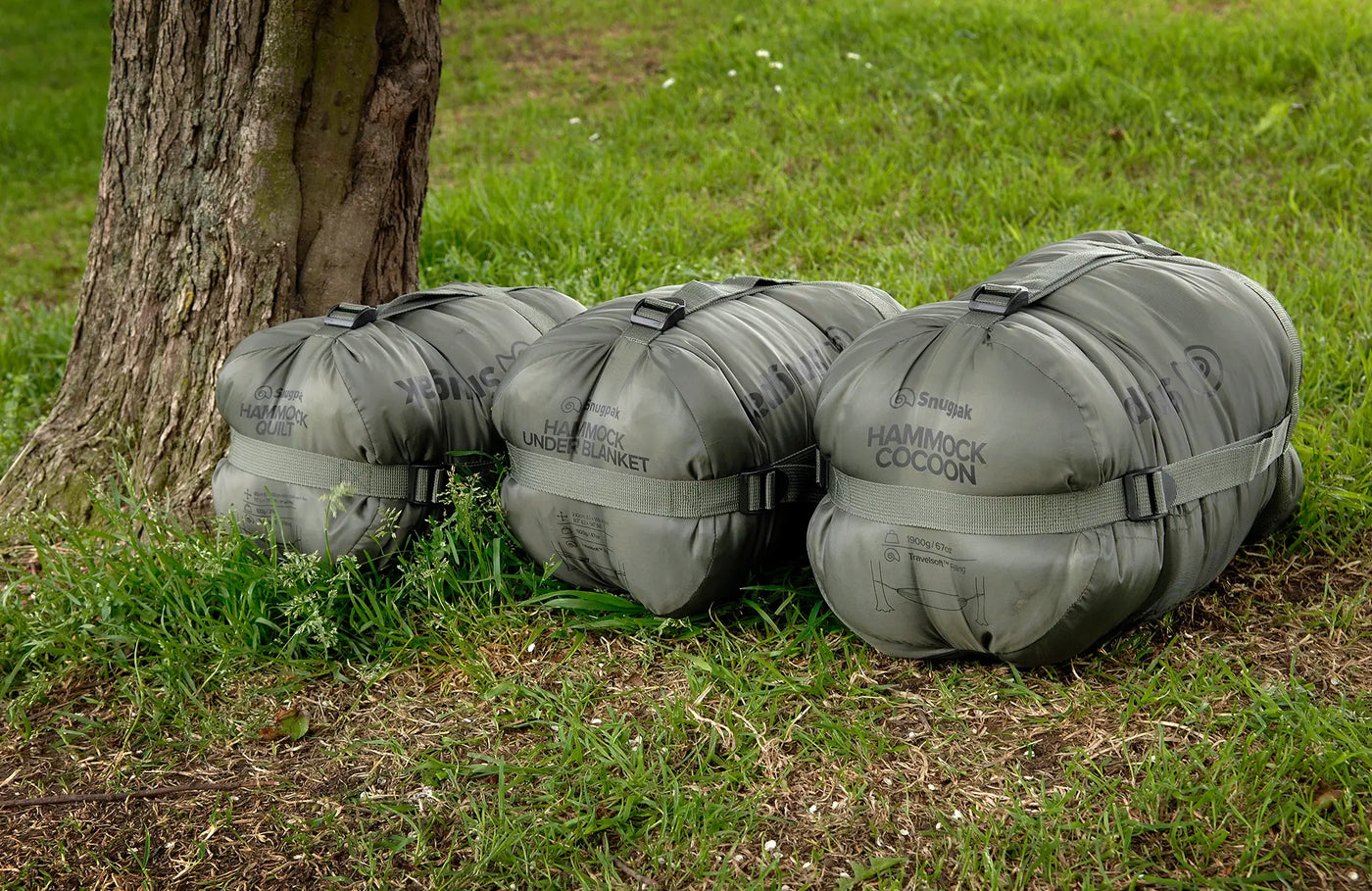Three olive green compressed Snugpak Hammock accessories together on the grass next to a tree trunk.