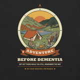 Close-up of the Adventure Before Dementia Camping T-Shirt print in Vintage Black: a couple by a campfire next to an orange tent beside a lake, with sheep, hills and a low sun inside a compass badge